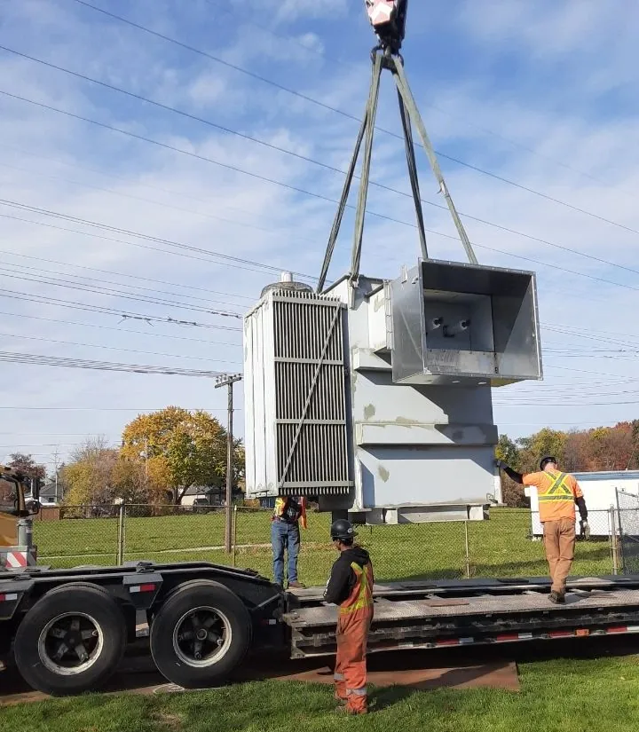 Power transformer lifted by crane onto a lowboy trailer while riggers guide it